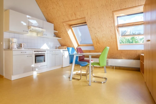 Helle Ferienwohnung mit Essecke, weißer Küchenzeile, Stühlen und Fensterfront. (KI generiert)