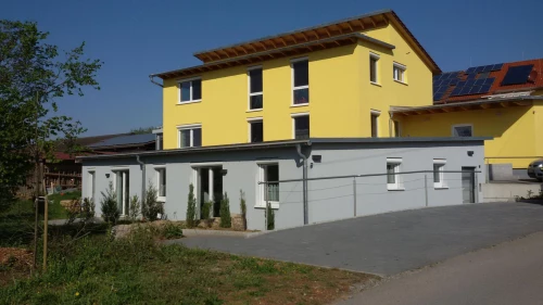 Ein modernes, gelbes Mehrfamilienhaus mit grauer Garage und Solaranlage auf dem Dach. (KI generiert)