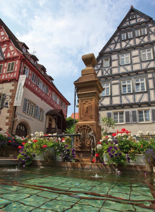 Ein Brunnen mit Blumen und zwei Fachwerkhäusern im Hintergrund. (KI generiert)