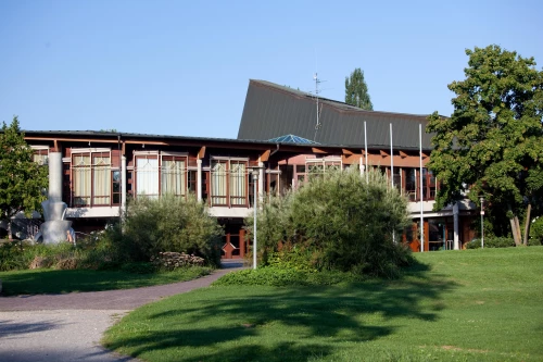 Ein großes, zweistöckiges Gebäude mit braunen Holzverkleidungen und großen Fenstern. Es hat ein flaches Dach und ist von Bäumen und Büschen umgeben. (KI generiert)