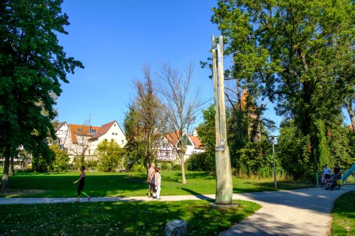 Park mit Menschen, Bäumen und Häusern im Hintergrund. Ein Kind spielt mit einem Ball. (KI generiert)