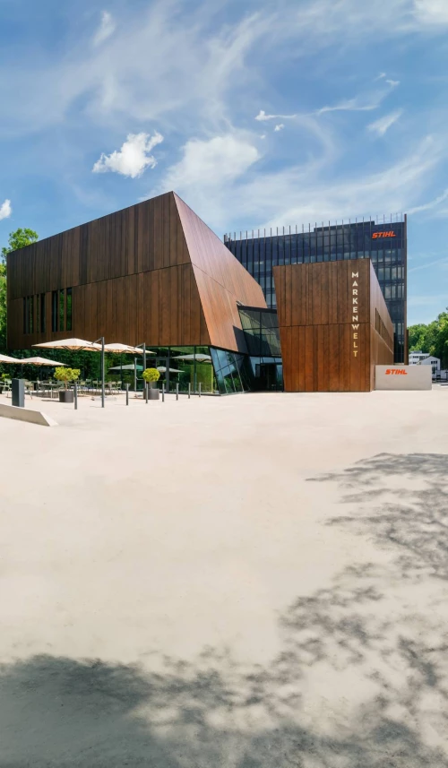 Moderne Markenwelt mit Holzfassade und Glasfronten, STIHL-Logo sichtbar, Außenbereich mit Tischen und Sonnenschirmen. (KI generiert)