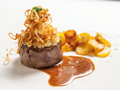 Ein Stück Fleisch mit Kräuterkruste, Zwiebelringen und Karamellsauce, garniert mit Zitrusfrüchten. (KI generiert)