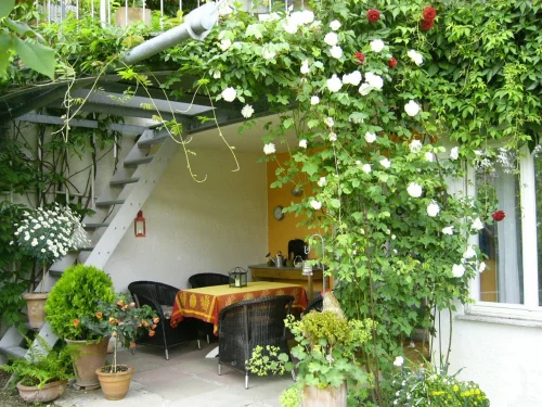 Ein überdachter Sitzbereich mit Tisch, Stühlen und vielen Pflanzen. Blühende Rosenranken hängen von der Decke. (KI generiert)