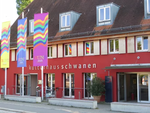 Rotes Gebäude mit mehreren Fenstern und bunten Fahnen, beschriftet 'kulturnausschwanen'. (KI generiert)