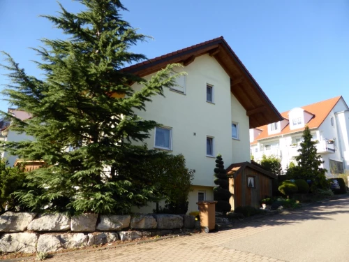 Ein gelbes Haus mit braunem Dach und einem kleinen Garten mit Bäumen und Sträuchern. (KI generiert)