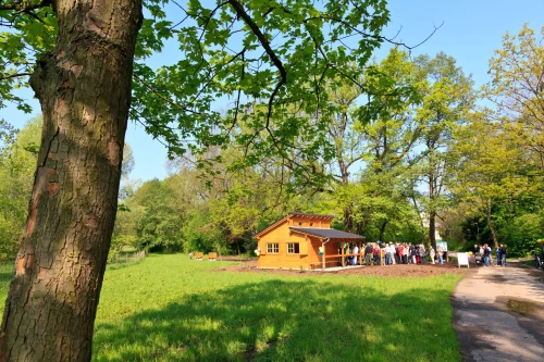 Eine Gruppe von Menschen steht vor einem kleinen, gelben Gebäude in einem Park mit grünen Bäumen und Gras. (KI generiert)
