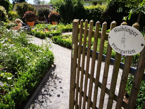 Ein Garten mit Blumenbeeten und einem Holzpfad, im Vordergrund ein Holzzaun mit einem Schild 'Waiblinger Pfarrgarten'. (KI generiert)