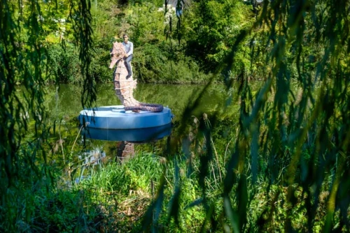 Eine Statue steht auf einer blauen Plattform in einem Teich mit grünen Pflanzen und Bäumen im Hintergrund. (KI generiert)