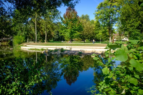 Ein ruhiger Park mit einem Teich, umgeben von Bäumen und Menschen, die spazieren gehen. (KI generiert)