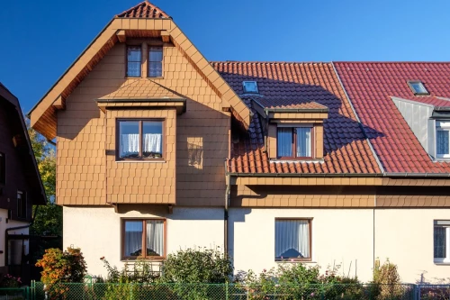 Zwei Häuser mit braunen Dachziegeln und weißen Wänden, umgeben von Büschen und Blumen. (KI generiert)