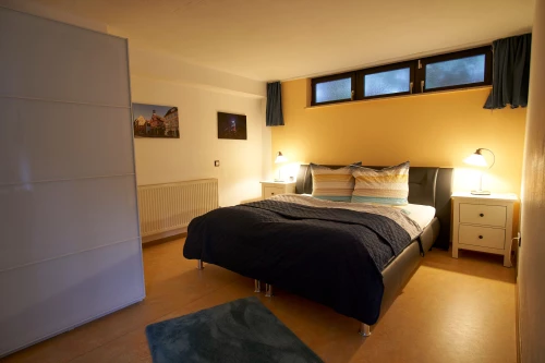 Ein Schlafzimmer mit einem Bett, zwei Nachttischen, einem Schrank und einem Heizkörper. Die Wände sind in hellen Farben gestrichen. (KI generiert)