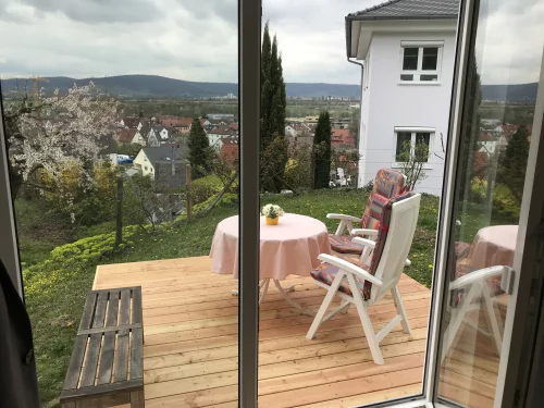 Terrasse mit Tischen, Stühlen und Blick auf ein Dorf und Hügel im Hintergrund. (KI generiert)