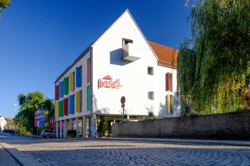 Ein weißes Gebäude mit farbigen Fensterläden und dem Schriftzug "Insel". (KI generiert)