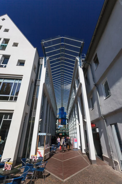 Eine überdachte Passage mit Glasdach, umgeben von Gebäuden mit Schaufenstern und Tischen mit Stühlen. (KI generiert)