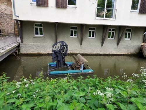 Ein Boot mit einem schwarzen Segel und einem braunen Segel auf einem Kanal, umgeben von grünen Pflanzen und einem Gebäude im Hintergrund. (KI generiert)