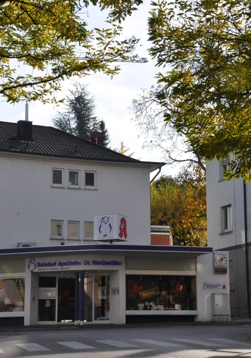 Apotheke mit blauem Eulen-Logo und Schaufensterauslage. Vor dem Gebäude ist eine Fußgängerampel. (KI generiert)