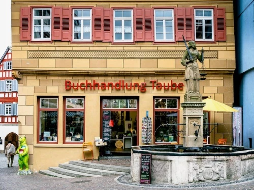 Buchhandlung mit gelber Fassade, roten Fensterläden und einer Statue davor. (KI generiert)