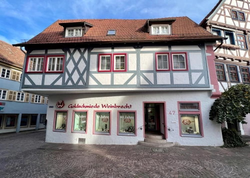 Ein zweistöckiges Fachwerkhaus mit weißem und grauem Fachwerk, rot gerahmten Fenstern und einem Ladenschild 'Goldschmiede Weinbrecht'. (KI generiert)