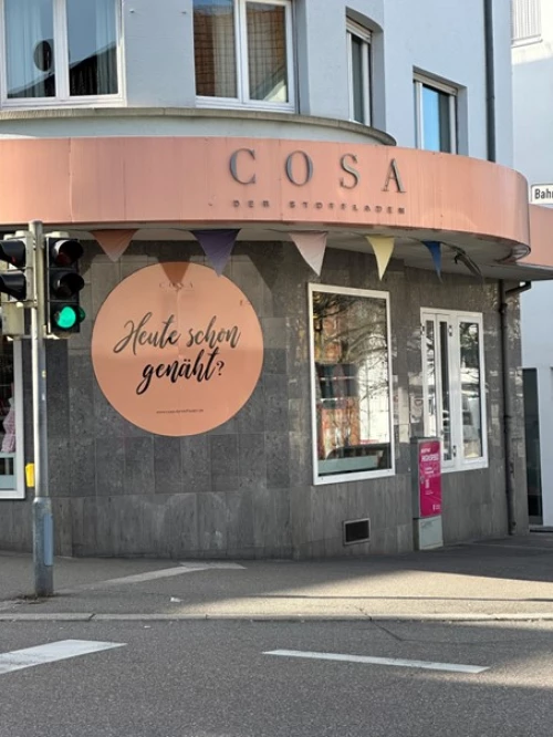 Ladenfront mit dem Namen COSA und einem Schild mit der Aufschrift Heute schon genäht? (KI generiert)