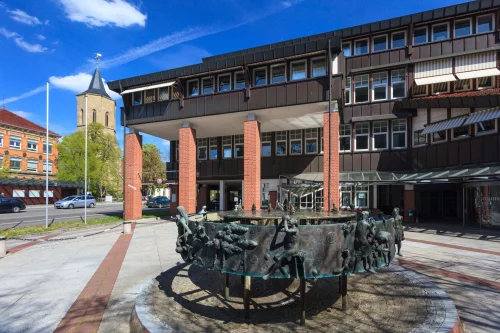 Straßenszene mit modernem Backsteingebäude, Pferdekutschen und Kirchturm im Hintergrund. (KI generiert)