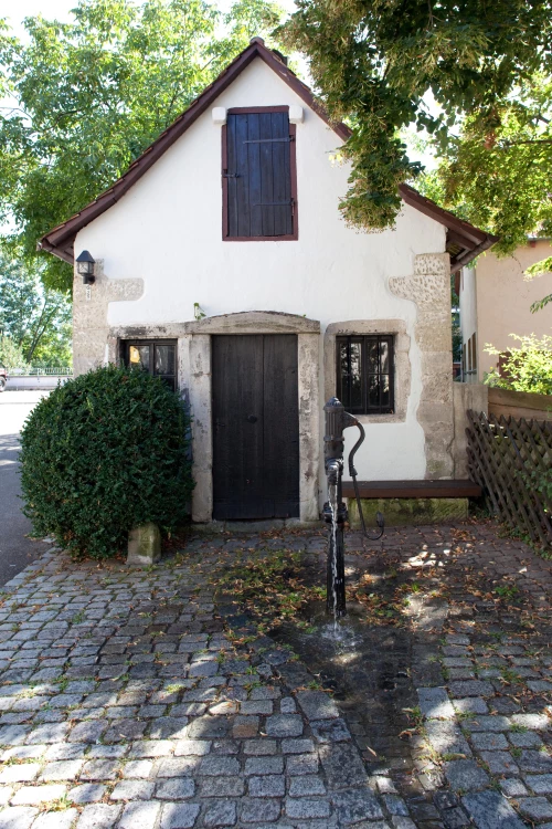 Altes Fachwerkhaus mit Steinfundament und Holzläden, umgeben von Bäumen und Kopfsteinpflaster. (KI generiert)