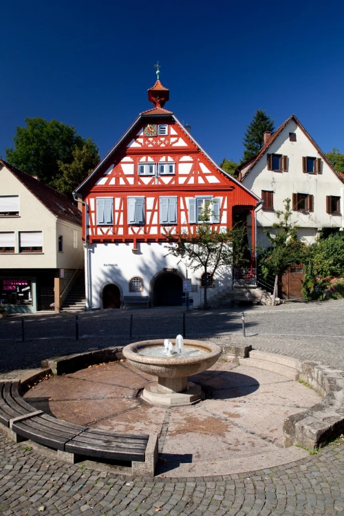 Rotes Rathaus mit Brunnen vor Fachwerkhäusern, sonniger Platz mit Sitzgelegenheiten. (KI generiert)