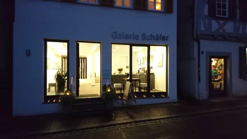 Galerie Schaefer, ein hell erleuchtetes Geschäft mit großen Fenstern, die Kunstwerke zeigen. Die Straße ist mit Pflastersteinen gepflastert und es gibt Pflanzen vor dem Geschäft. (KI generiert)