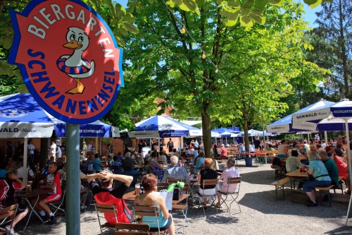 Biergarten mit vielen Menschen, Tischen und Schirmen unter Bäumen. Ein Schild mit einem Vogel und der Aufschrift 'Biergarten Schwabenhüsel' ist prominent zu sehen. (KI generiert)