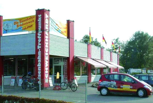 Ein Gebäude mit dem Namen "Die Eisenbahn-Erlebniswelt mit Pfiff" und mehreren Flaggen davor. Ein rotes Auto ist geparkt. (KI generiert)