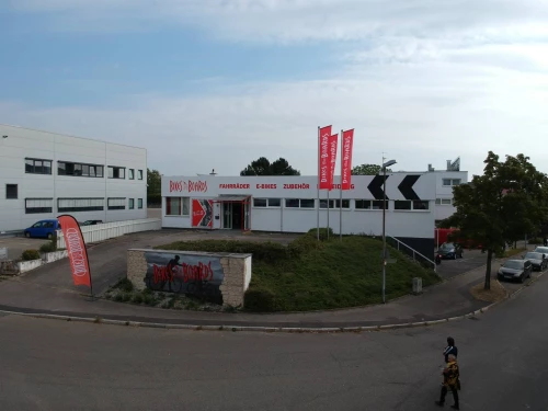 Ein Gebäude mit dem Namen Bikes & Boards, das Fahrräder, E-Bikes und Zubehör anbietet. Vor dem Gebäude sind mehrere Fahnen und Schilder. (KI generiert)