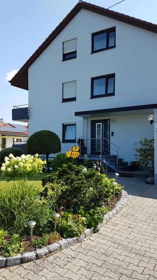 Zweistöckiges Haus mit weißer Fassade, Garten mit Blumen und gepflastertem Weg. Bienen-Deko im Garten. (KI generiert)