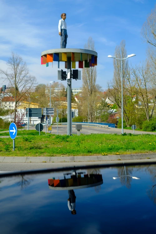 Eine Statue eines Mannes steht auf einer bunten, kreisförmigen Plattform auf einem hohen Pfahl. Die Plattform spiegelt sich im Wasser darunter. (KI generiert)