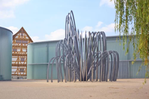 Moderne Metallskulptur vor einem Gebäude mit Fachwerk und modernen Elementen. (KI generiert)
