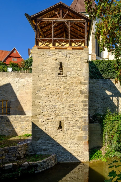 Alter steinerner Turm mit Holzdach und zwei Schießscharten, umgeben von Grün und Wasser. (KI generiert)