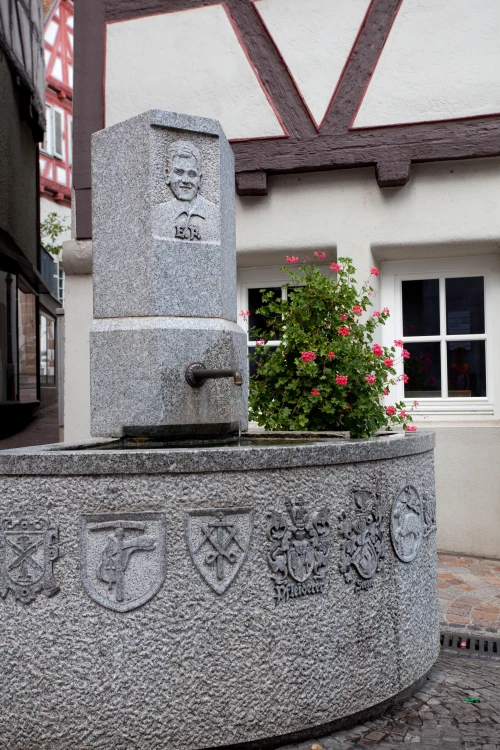 Steinbrunnen mit Gravuren und Blumen, vor einem Fachwerkhaus. (KI generiert)