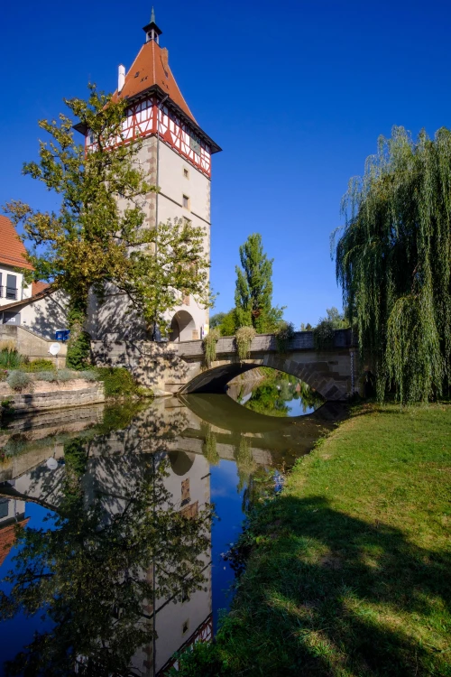 Ein Turm mit rotem Dach und weißer Fassade, umgeben von Bäumen und einem Teich mit einer Brücke. (KI generiert)