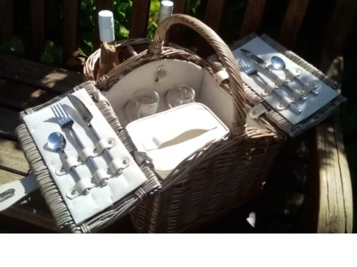 Ein Picknickkorb mit Besteck, Gläsern und einem Kühlbehälter auf einem Holztisch. (KI generiert)