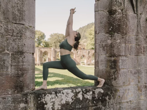 Person in grüner Sportkleidung macht Yoga-Pose auf einer Wiese vor alten Ruinen. (KI generiert)